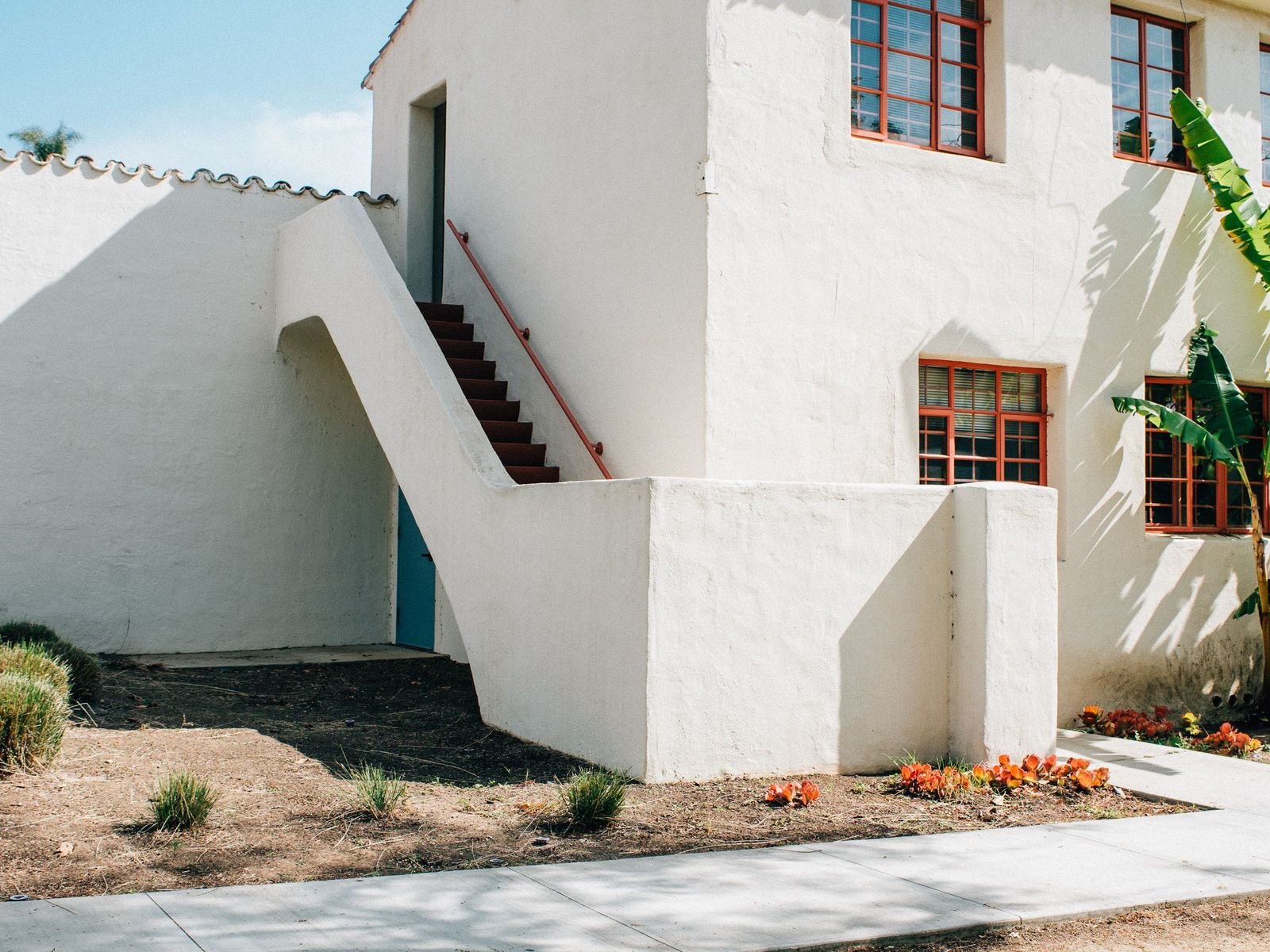 White stucco building where the wall meets bare earth, with dry grass tufts and low planting set against the base and a concrete walkway alongside