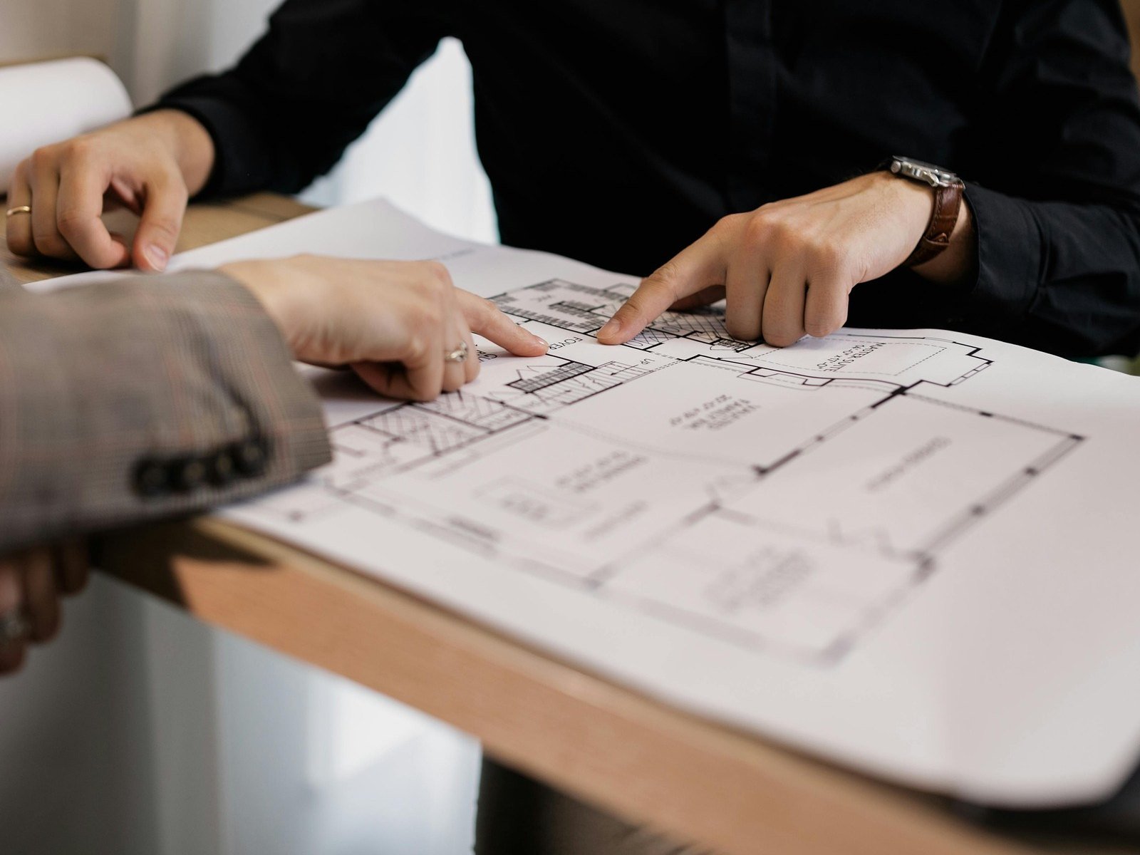 Two people leaning over a set of architectural floor plans on a desk, both pointing at the same detail on the drawing